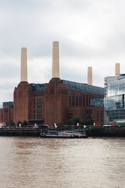 A large, historic brick building with four tall, white smokestacks rises above the River Thames in London. The building's facade features vertical windows and decorative brickwork, illuminated by natu