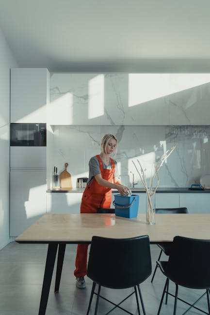 A woman in a grey top and red apron is performing domestic cleaning in a modern kitchen, which features a large wooden dining table with black chairs, and a sleek white marble backsplash. She is placing items into a blue container, possibly preparing for deep cleaning or sanitisation. The kitchen is illuminated by natural sunlight streaming through a large window, casting bright rectangular light patches on the wall and counter, highlighting the clean and tidy surfaces. Visible on the countertop are a clear glass vase with decorative branches, a cutting board, and some cleaning products. The overall setting demonstrates a hygienic, well-maintained space with polished surfaces, including ceramic tiles and stainless steel fixtures, emphasizing surface cleaning and hygiene focused on domestic cleaning services provided by Battersea Cleaner.